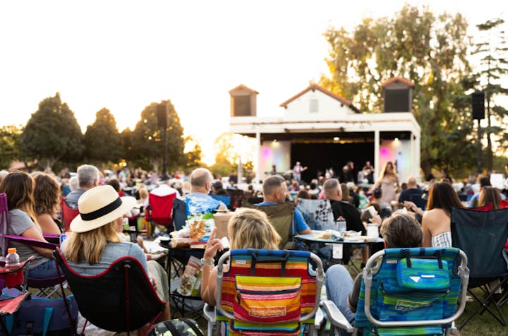 Outdoor event with people sitting on chairs enjoying a performance on stage at dusk.