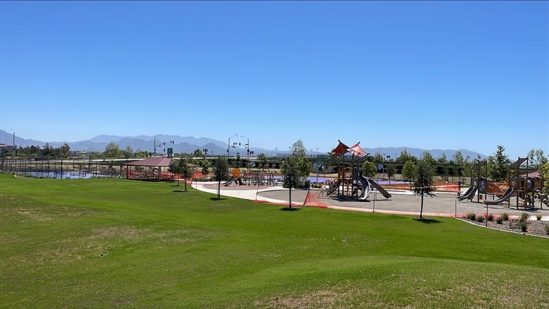 A sunny park scene featuring a playground, grassy areas, and mountains in the background.