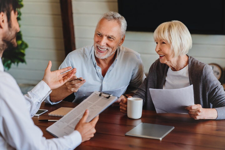 A smiling older couple discussing paperwork with a person holding a clipboard at a table.