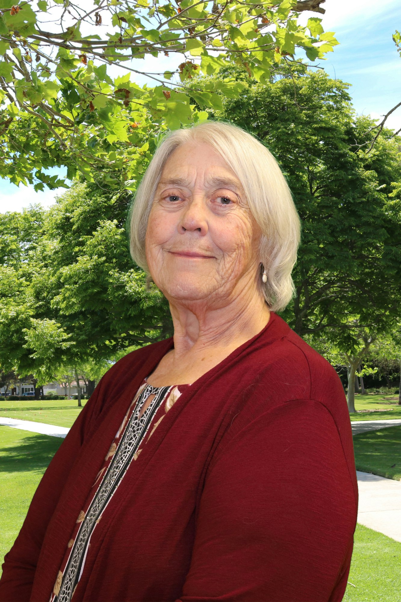 An elderly woman with white hair wearing a burgundy outfit stands outdoors with green trees and clear skies in the background.