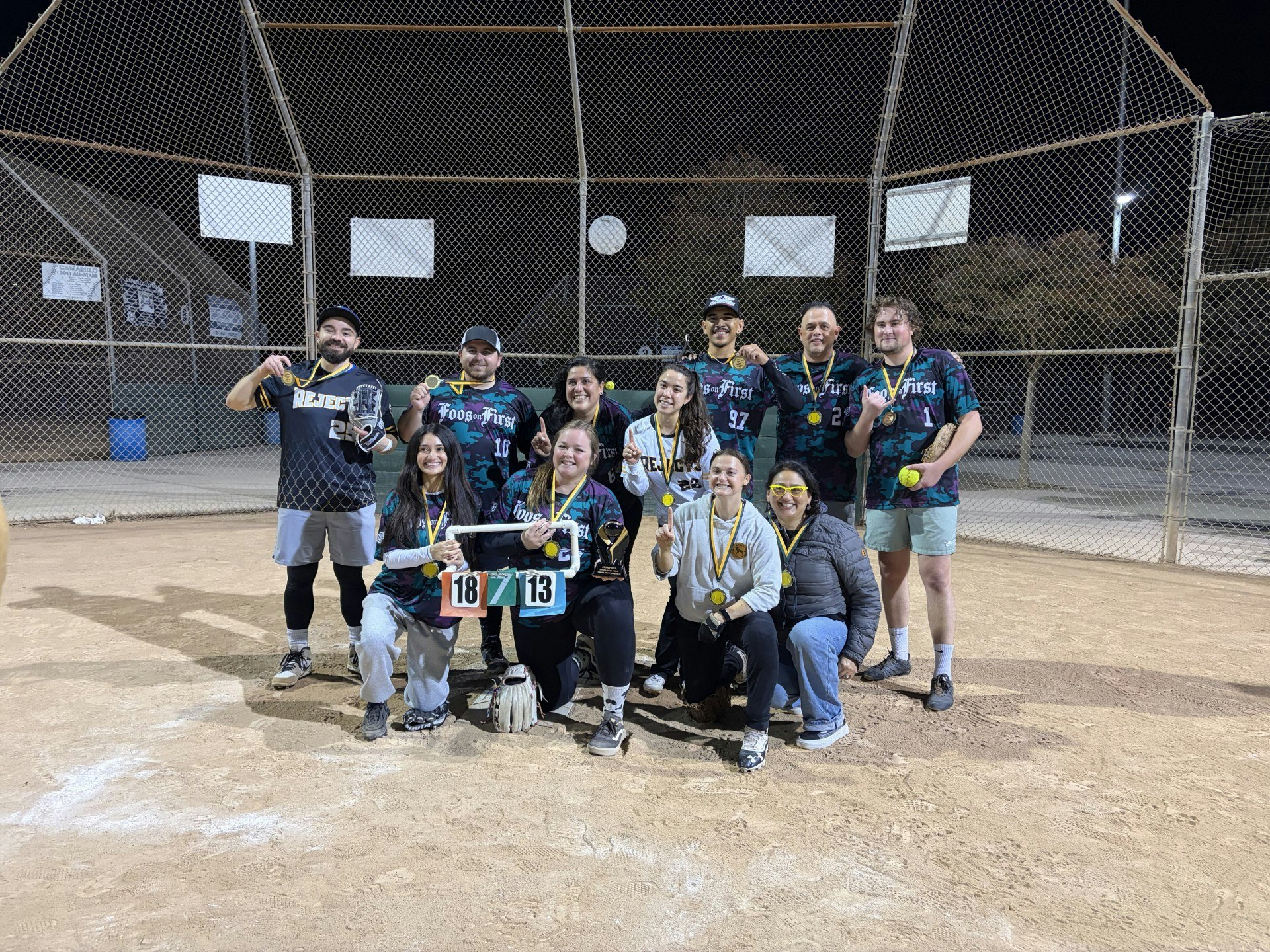 A softball team poses with trophies and medals after winning a game, celebrating their victory at night on the field.