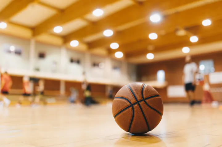 A basketball on a shiny gym floor with players in the background.