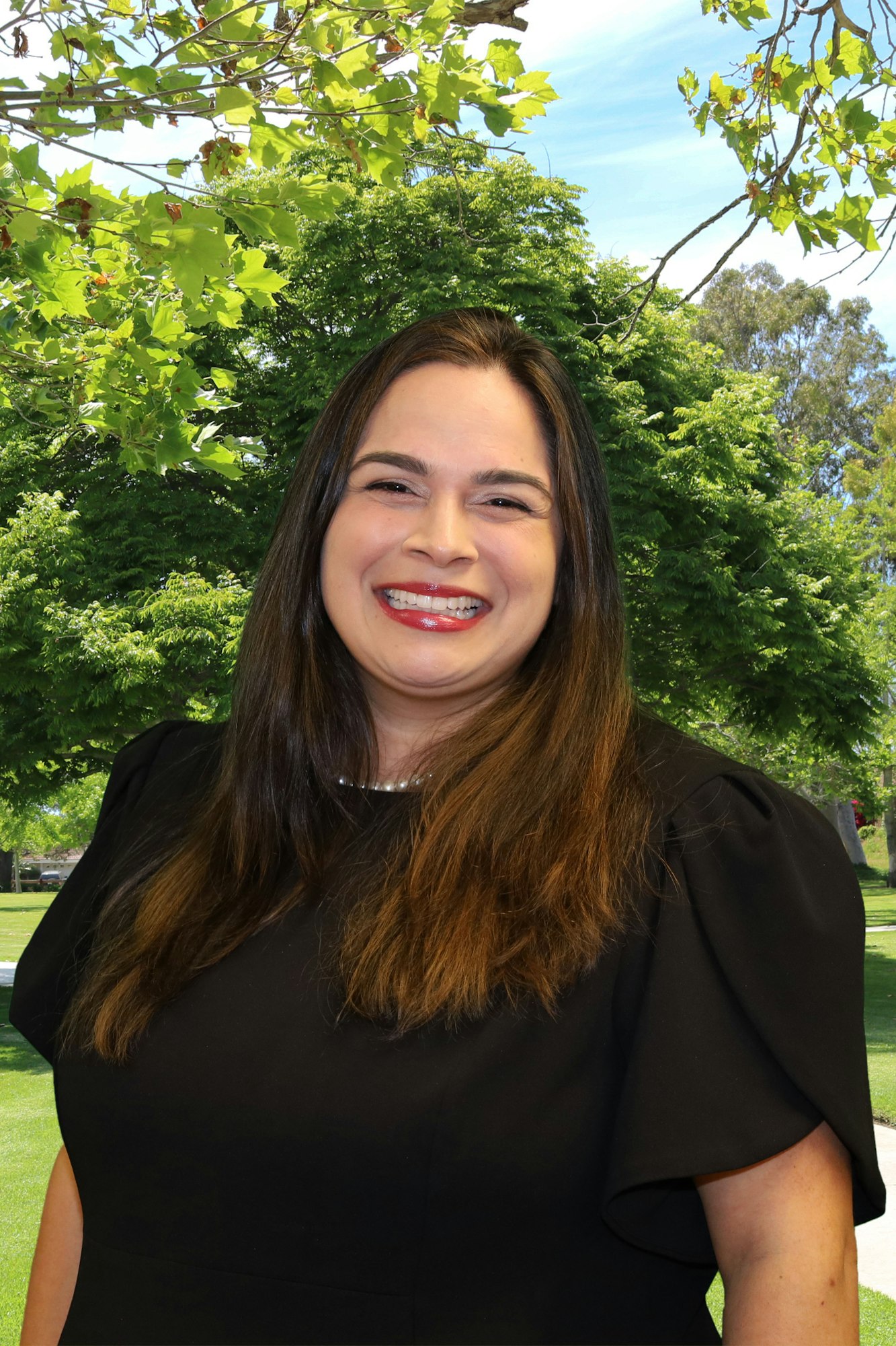 A smiling individual with long hair and a black dress, standing outdoors with green trees in the background.