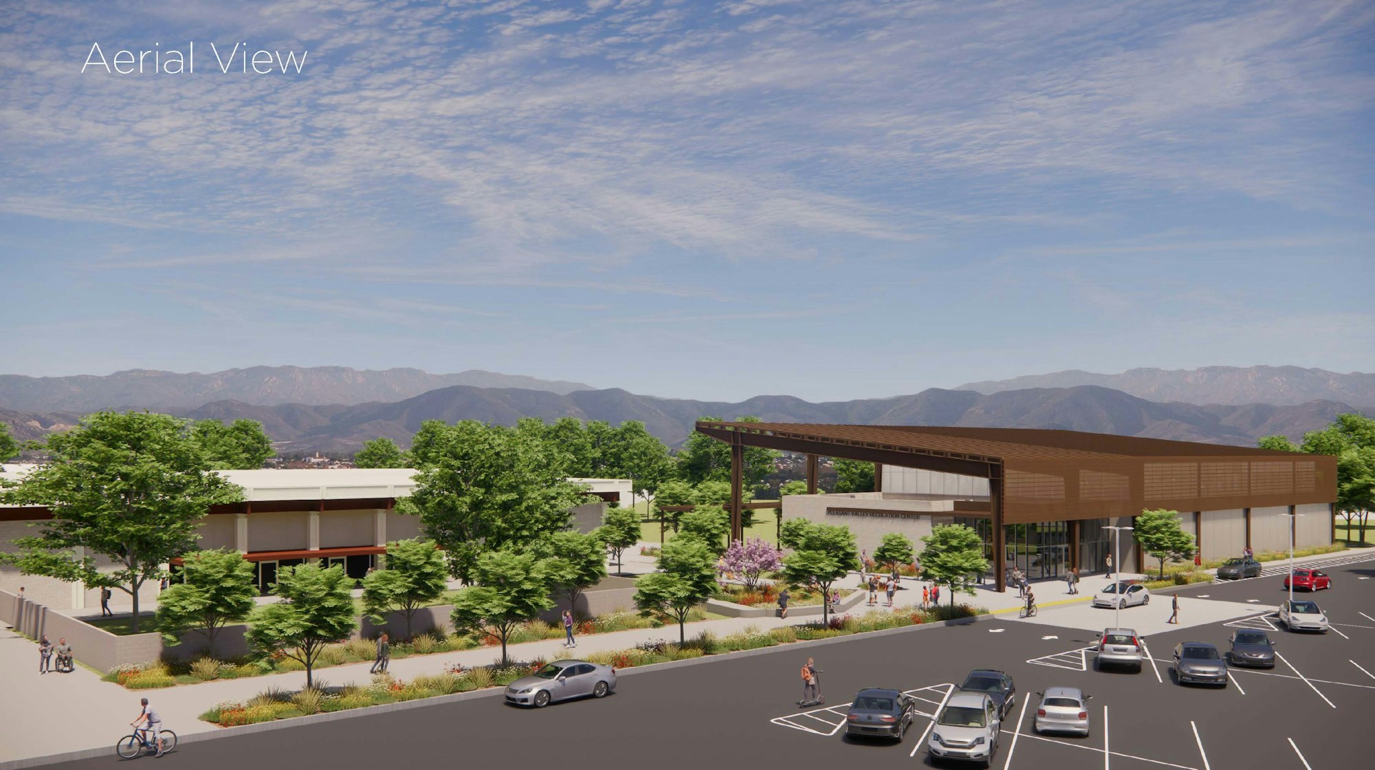 An aerial view of a modern building surrounded by greenery, mountains in the background, with parking and pathways.