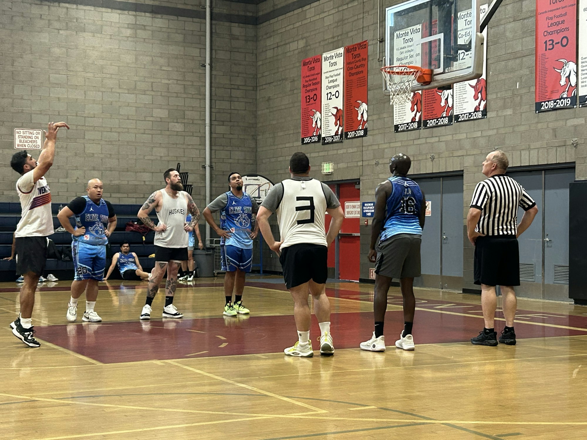 A basketball game in progress with players on court and a referee, in a gym with banners on the wall.