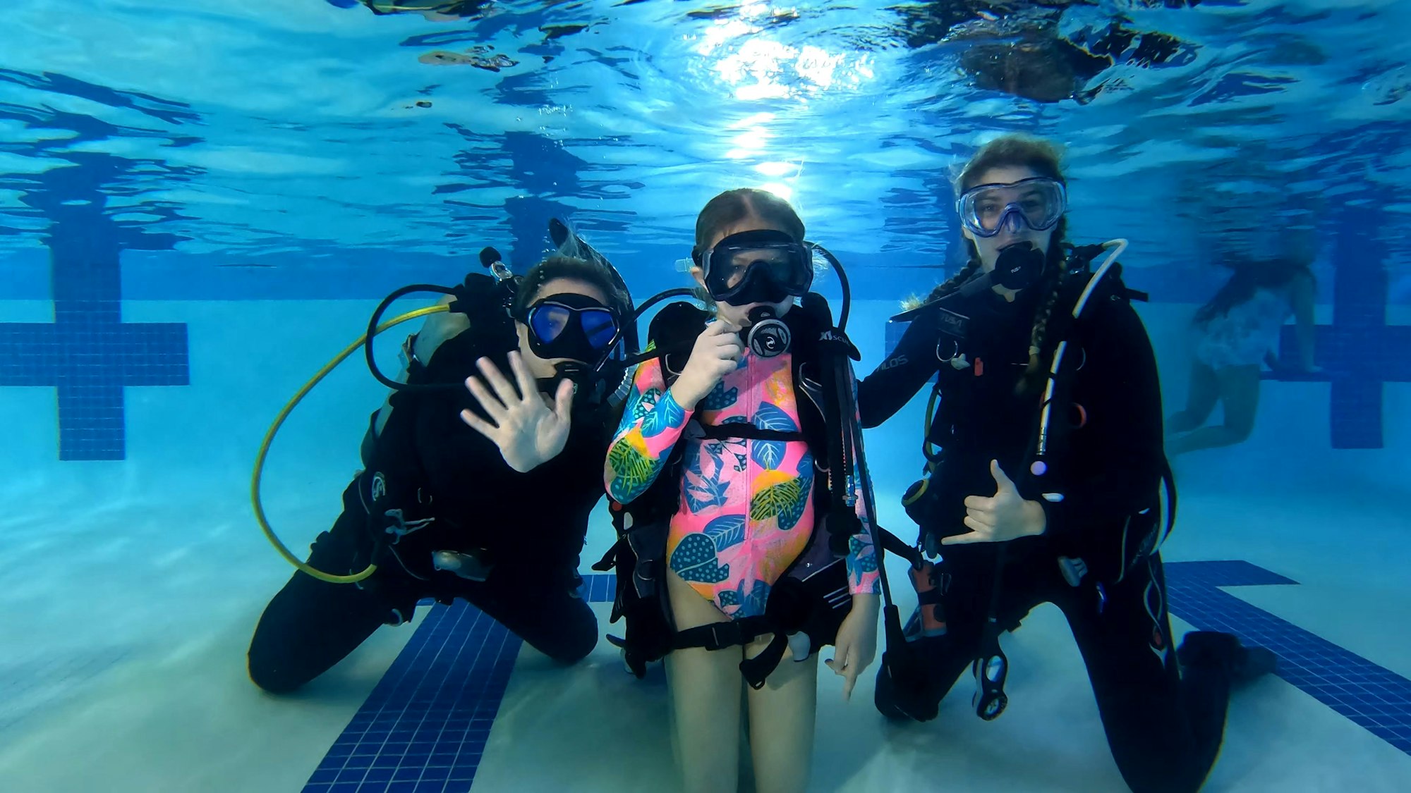 Three scuba divers in a pool, all wearing diving gear and giving hand signals, with a clear underwater view.