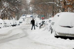 A person walking down a snowy street lined with snow-covered cars.