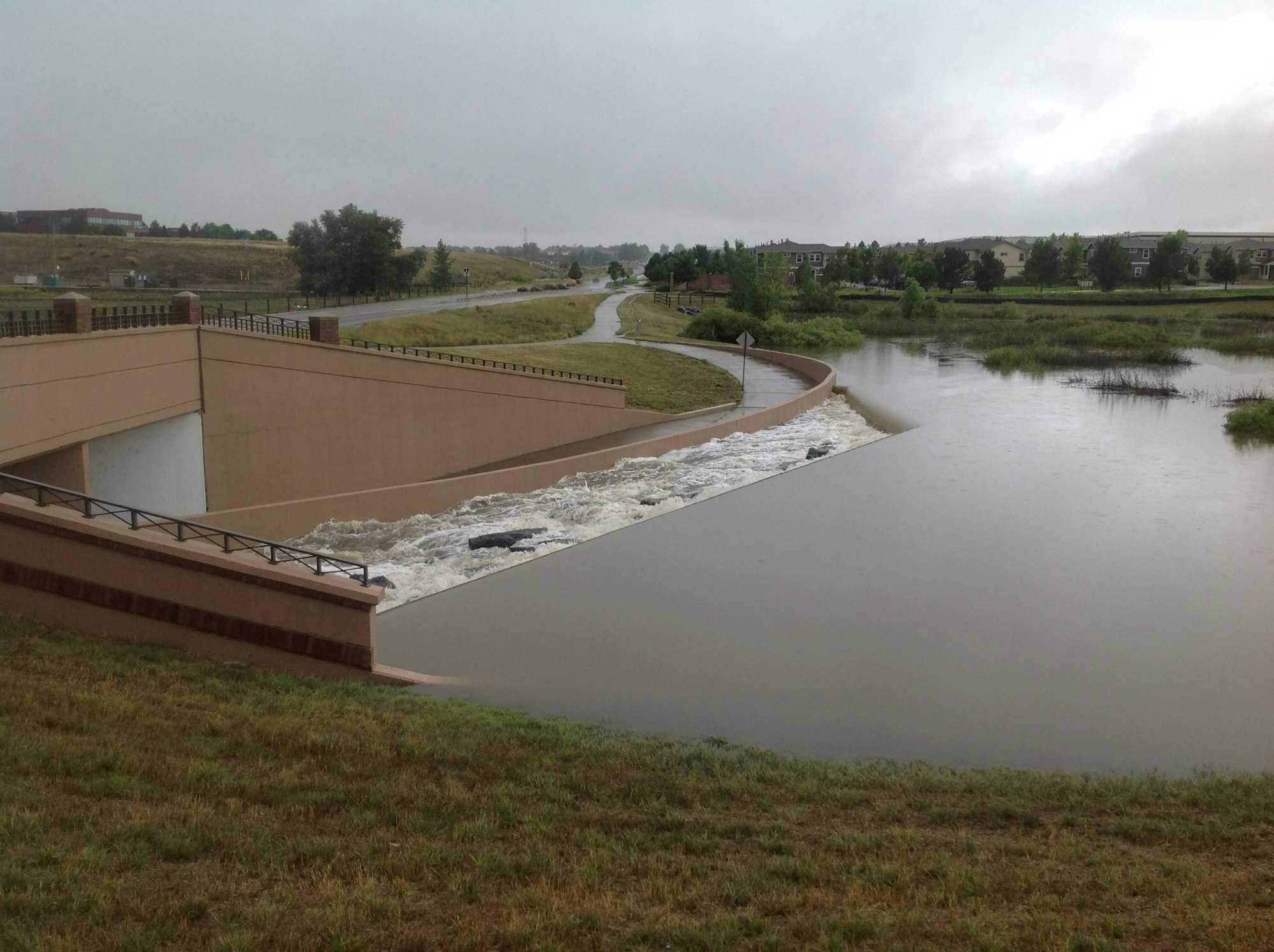 A waterway with a spillway, overcast sky, and surrounding greenery, potentially in a suburban area.