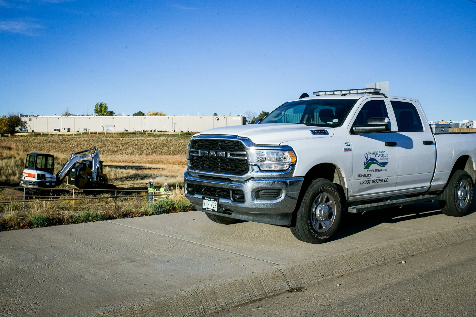 A white RAM pickup truck and a mini excavator with a worker nearby; industrial setting.