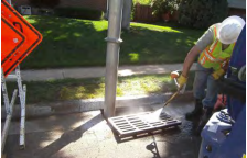 A worker in a high-visibility vest is using a tool near a storm drain, with a detour traffic sign and ladder in the background.