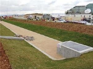 A pathway ends abruptly above a storm drain, with rocks scattered alongside and buildings in the distance.
