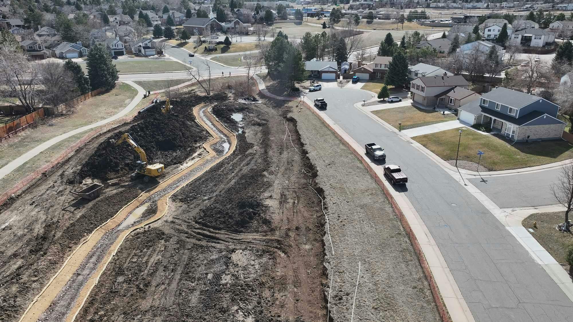 Residential area with construction site, showing machinery, dirt mounds, and a winding path near houses and streets.