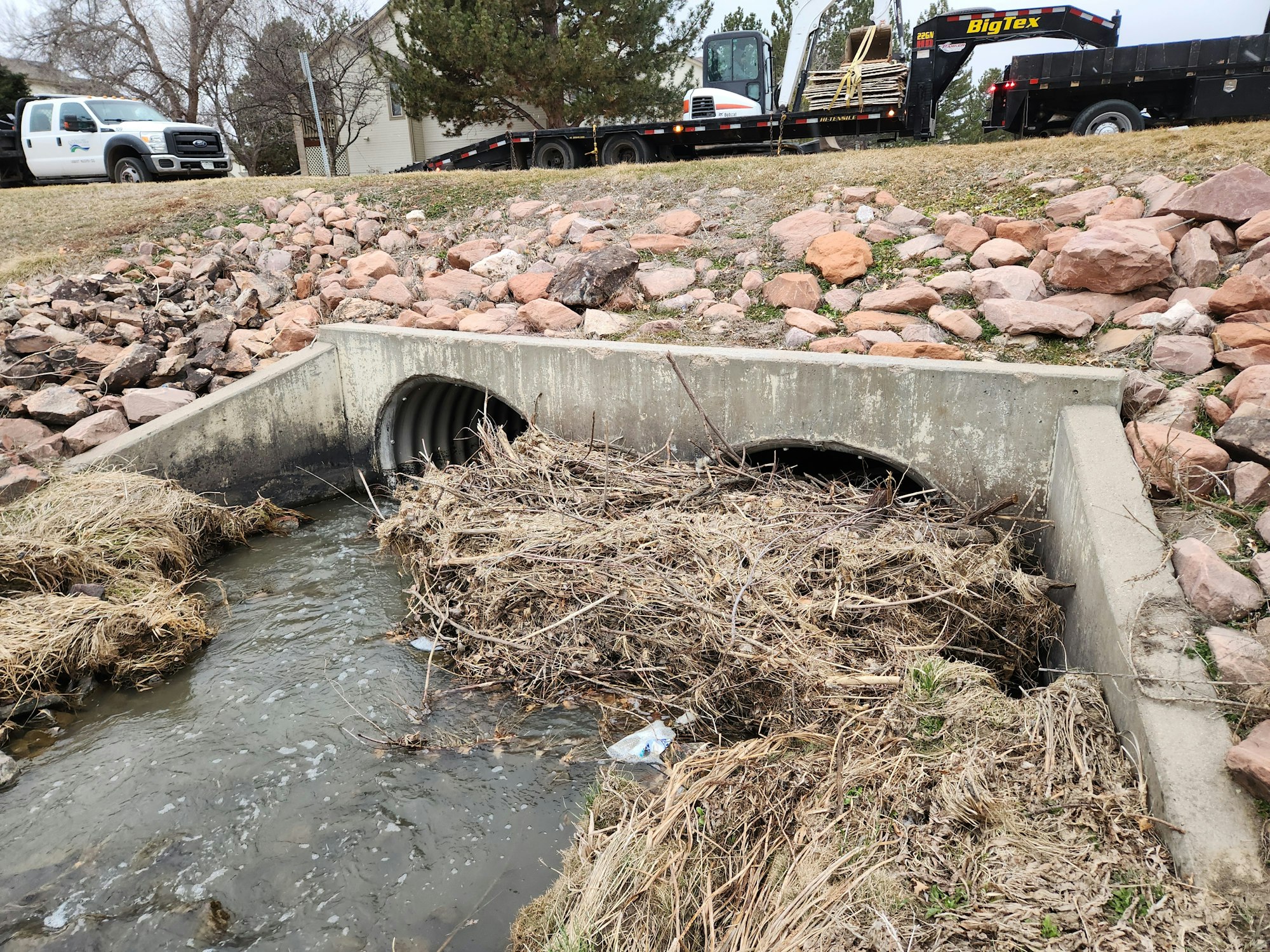 A drainage culvert with debris buildup, water flow, rocks, with vehicles and a trailer in the background.