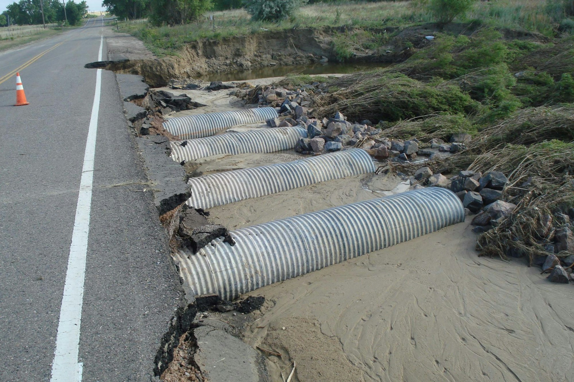 A damaged road with exposed, bent culvert pipes and erosion at the edge.