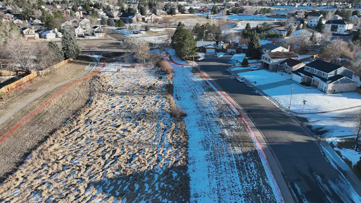 Suburban neighborhood with snow patches, road, houses, trees, and grassy area.