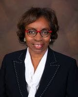 A person wearing glasses, a dark suit, and a white blouse poses against a brown backdrop.