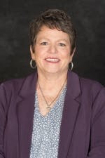 A smiling woman with short hair, wearing a purple blazer over a patterned blouse, with earrings and a necklace, against a dark background.
