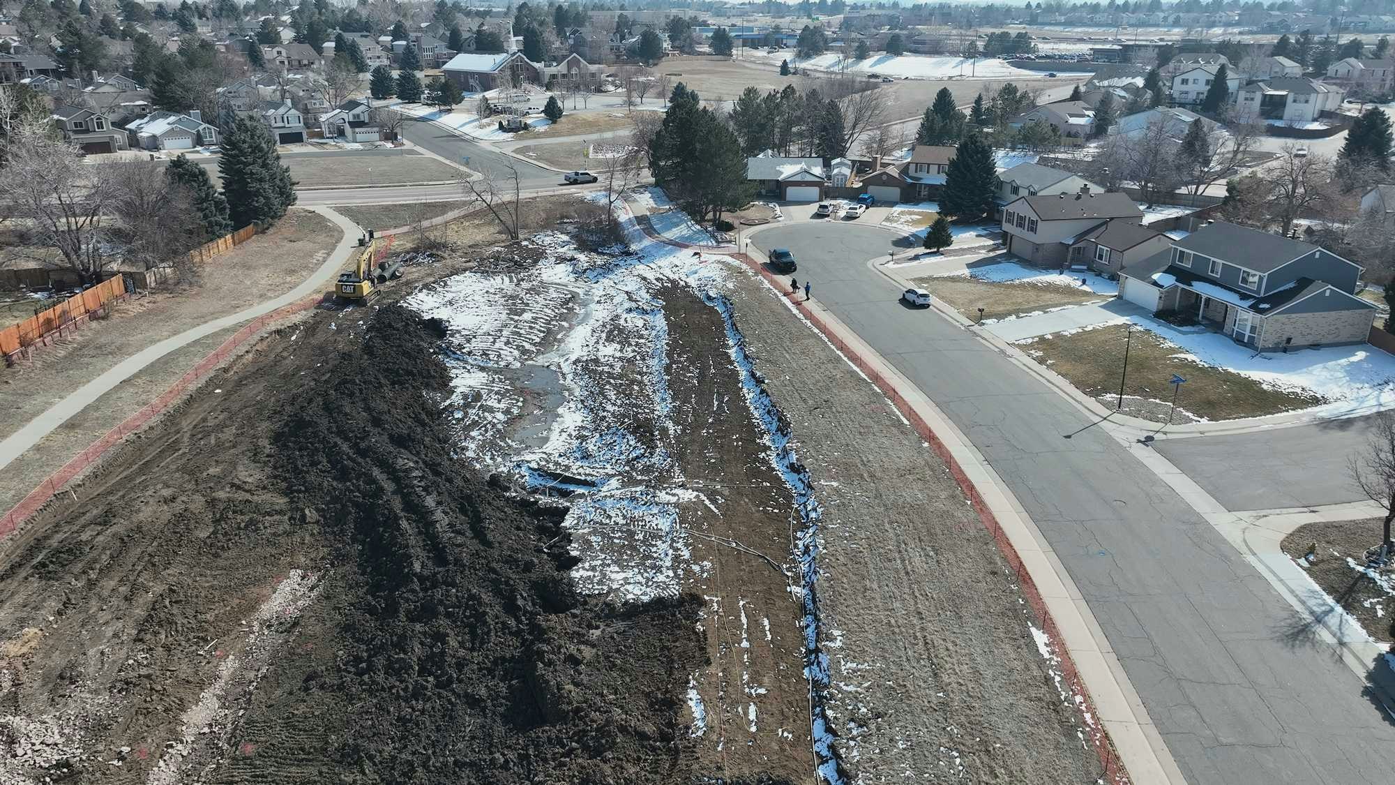 Suburban area with a construction site, snow patches, houses, and parked cars. A pathway runs alongside the site.