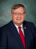A smiling man in a blue suit with a flag lapel pin, against a textured backdrop.