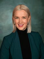 A smiling woman with blonde hair and earrings, wearing a black top and a teal blazer.