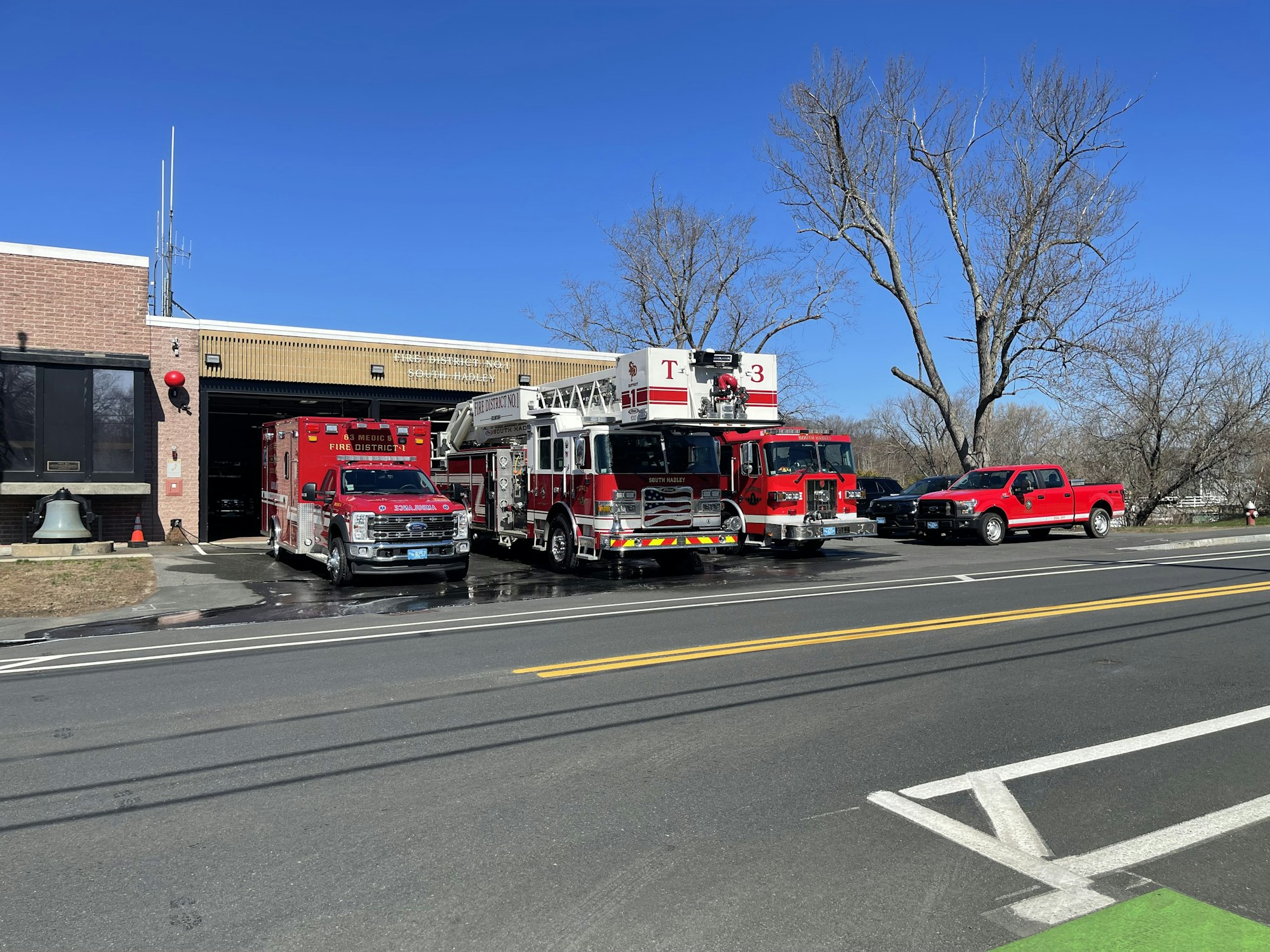 The image shows a fire station with fire trucks, an ambulance, and a red pickup parked outside under a clear blue sky.