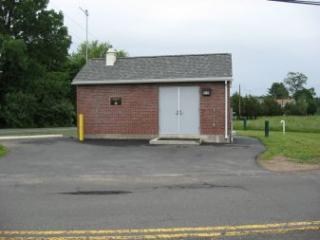 A small brick utility building with a gray door, surrounded by pavement and some greenery.