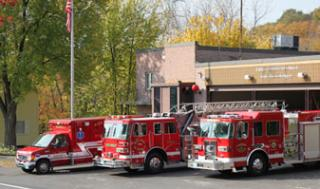 Fire station with red fire trucks and an emergency vehicle parked outside, surrounded by trees and an American flag.