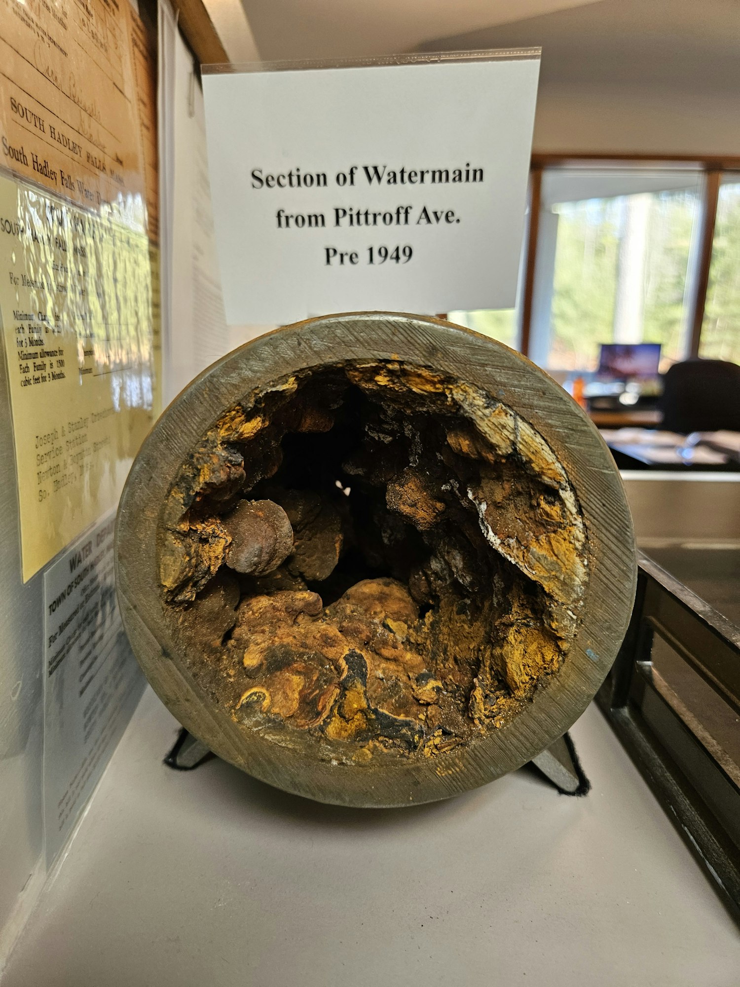 A section of a rusty water 4" Watermain pipe from Pittroff Ave., dated pre-1949, displayed on a stand with a sign.