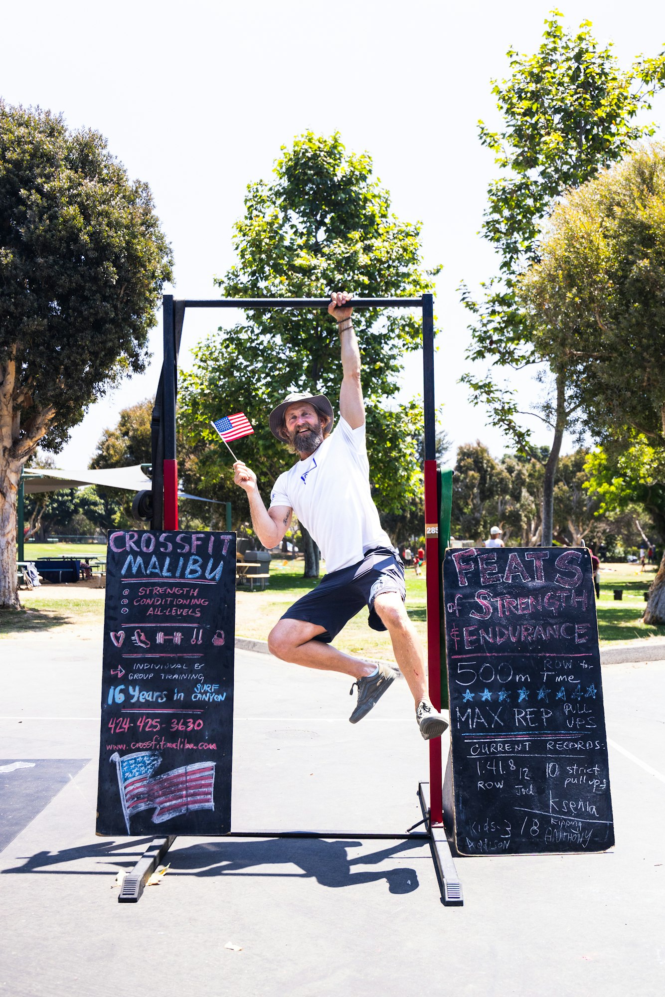 Man hanging on a pull-up bar at an outdoor event, holding a small American flag, with chalkboards about fitness challenges.