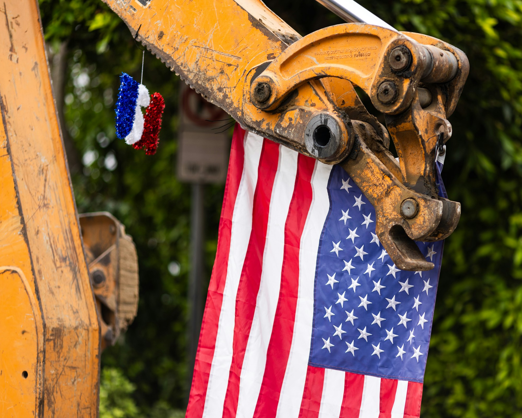 A construction vehicle arm holds a U.S. flag with a small red, white, and blue decoration hanging.