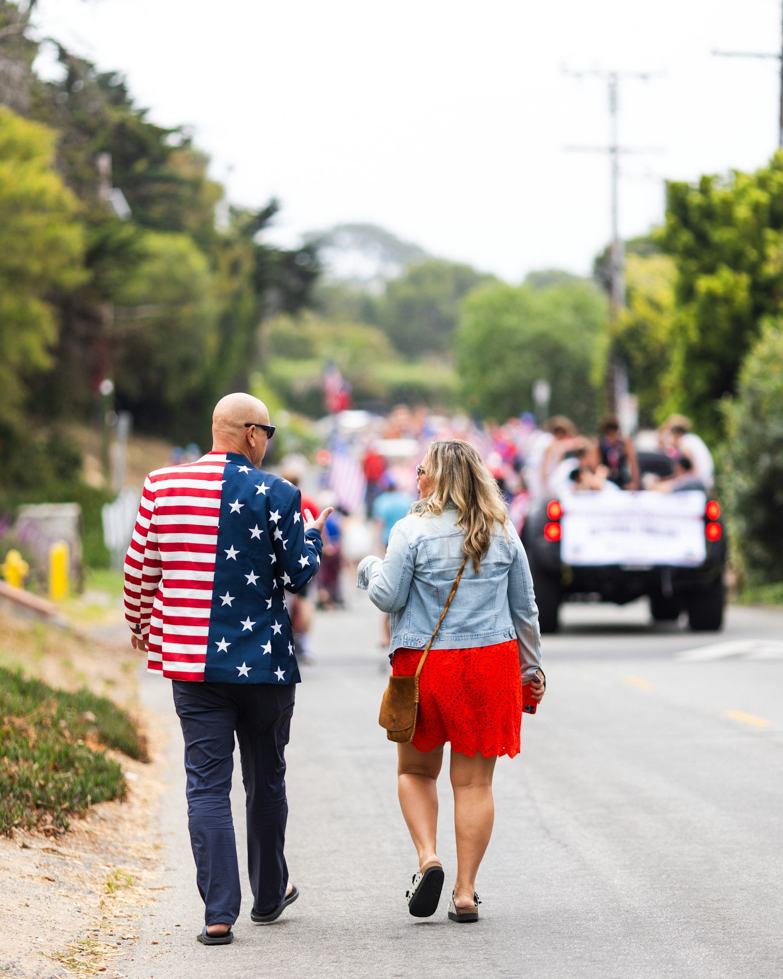 A man in a stars and stripes blazer and a woman walk down a street towards a parade.