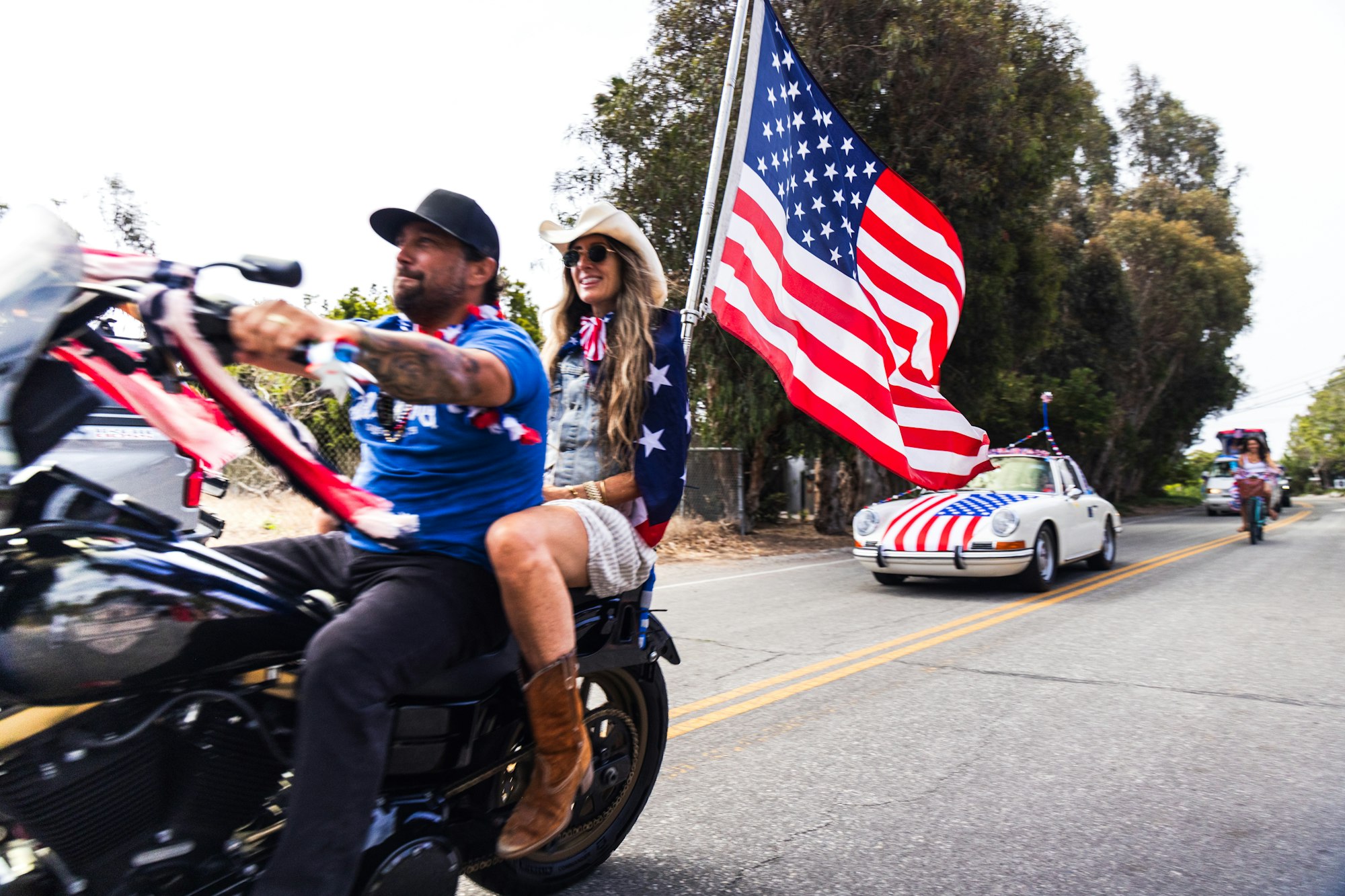 Motorcycle with two riders waving an American flag, followed by a car with flag decorations on a road.