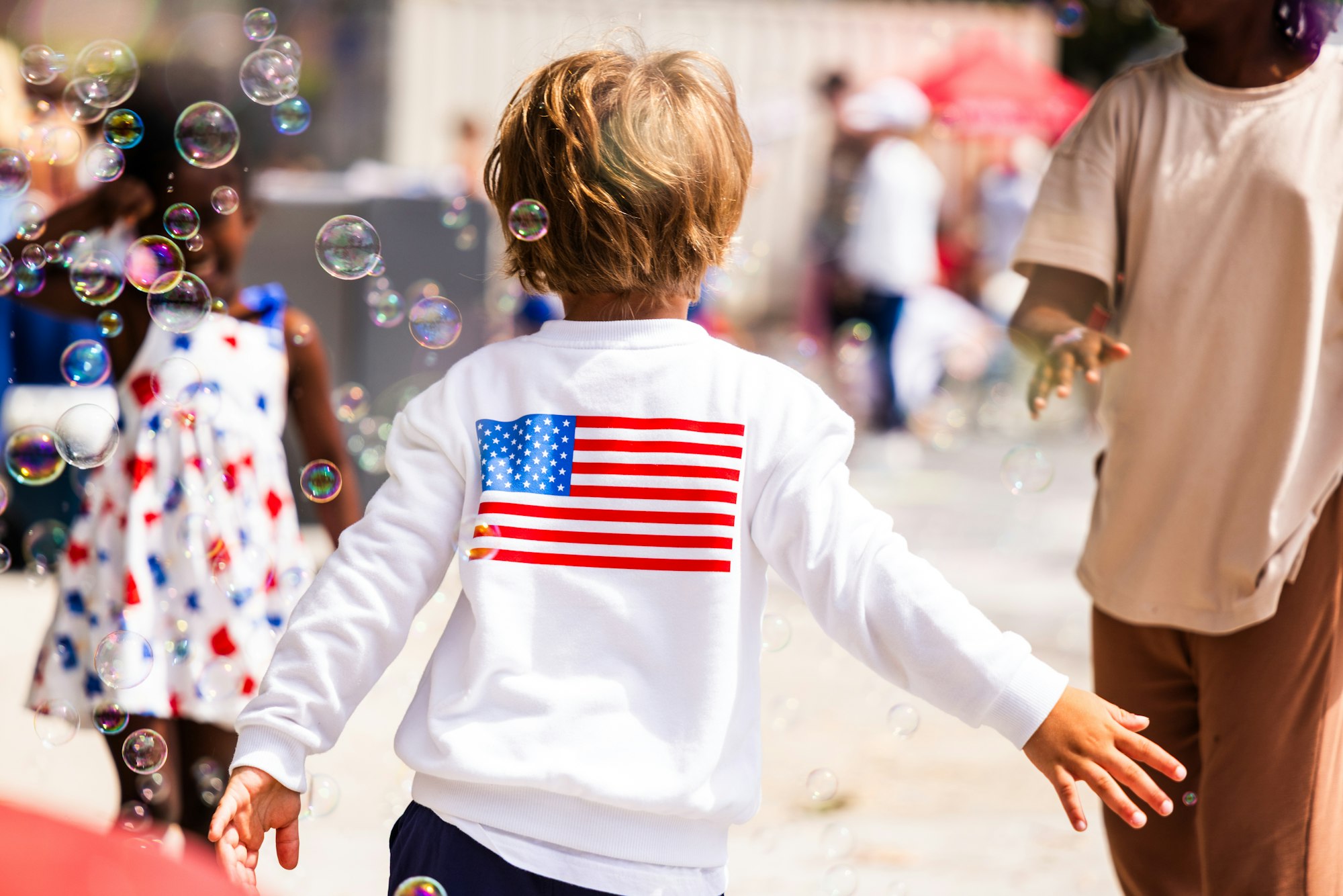 Child in a white sweatshirt with an American flag, playing with bubbles outdoors.