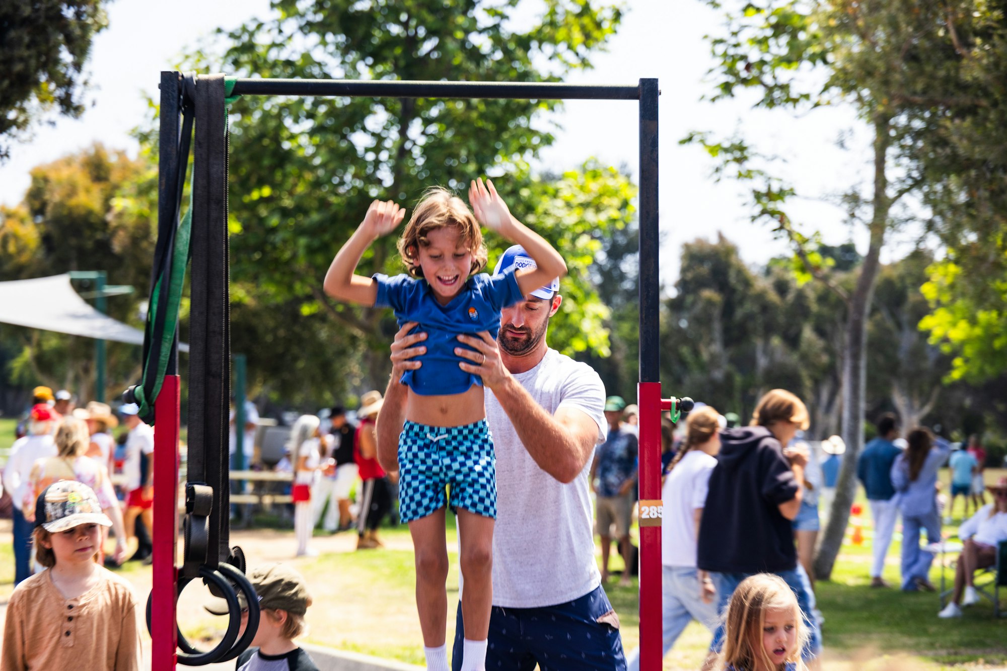 A man helps a child exercise on a pull-up bar at an outdoor event with people in the background.