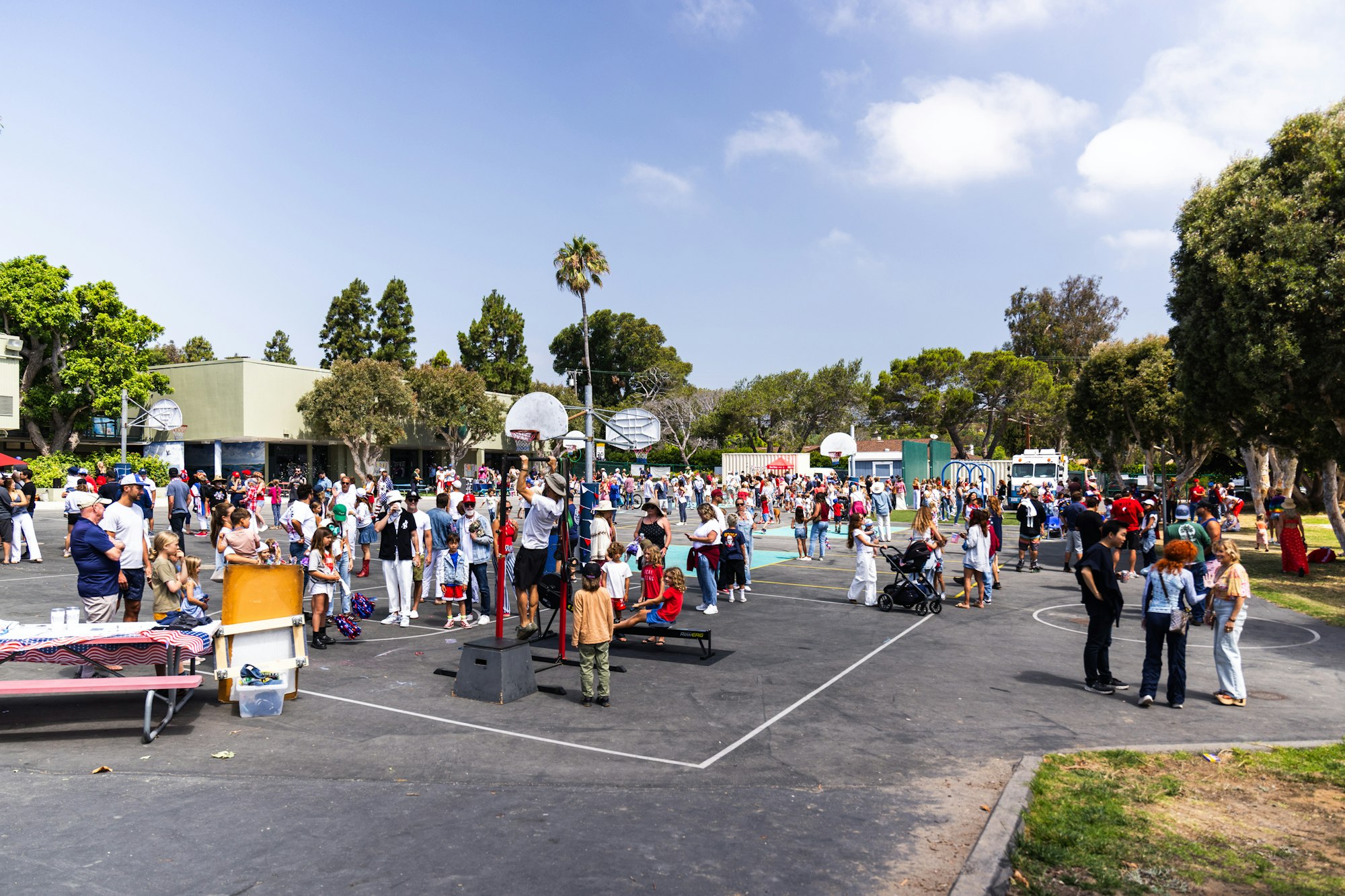 A crowded outdoor event with people gathered around basketball courts, strollers, and trees under a clear sky.