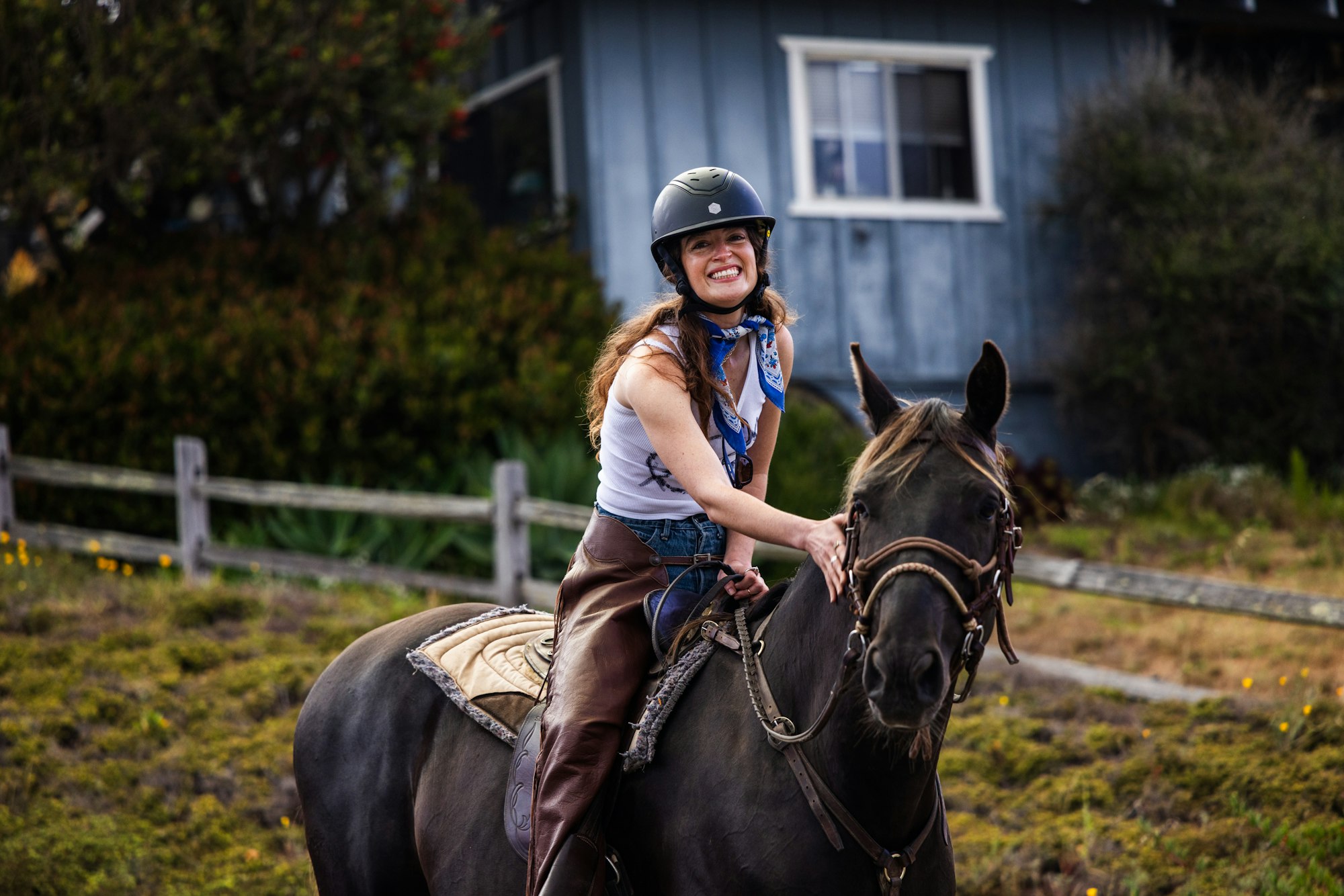 A smiling person wearing a helmet and scarf rides a horse near a blue house and a wooden fence.