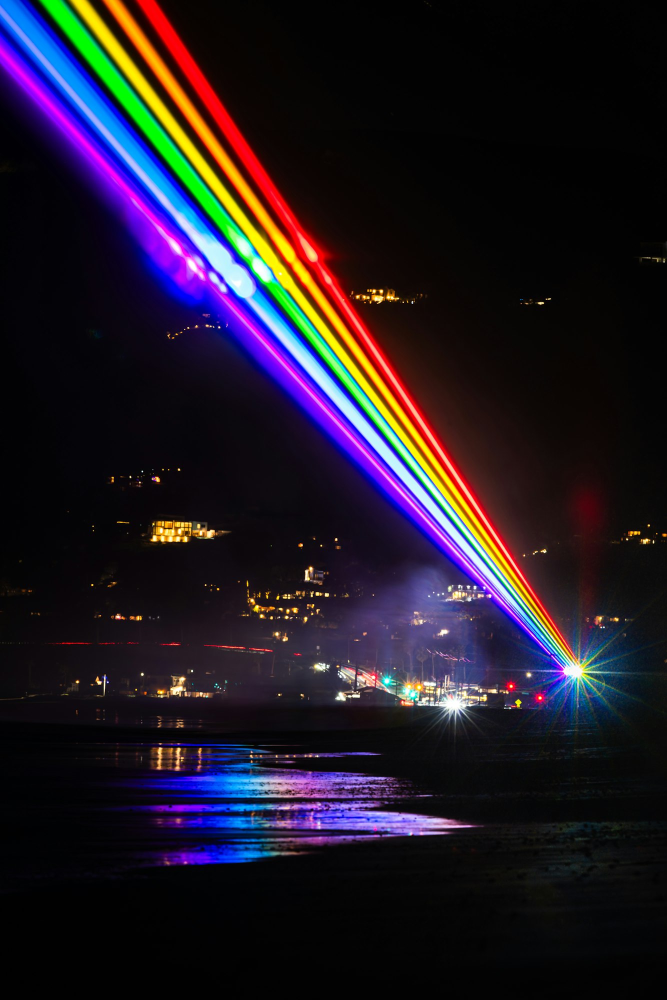 A vibrant display of rainbow beams lights up the night sky, reflecting off the water and illuminating nearby buildings.