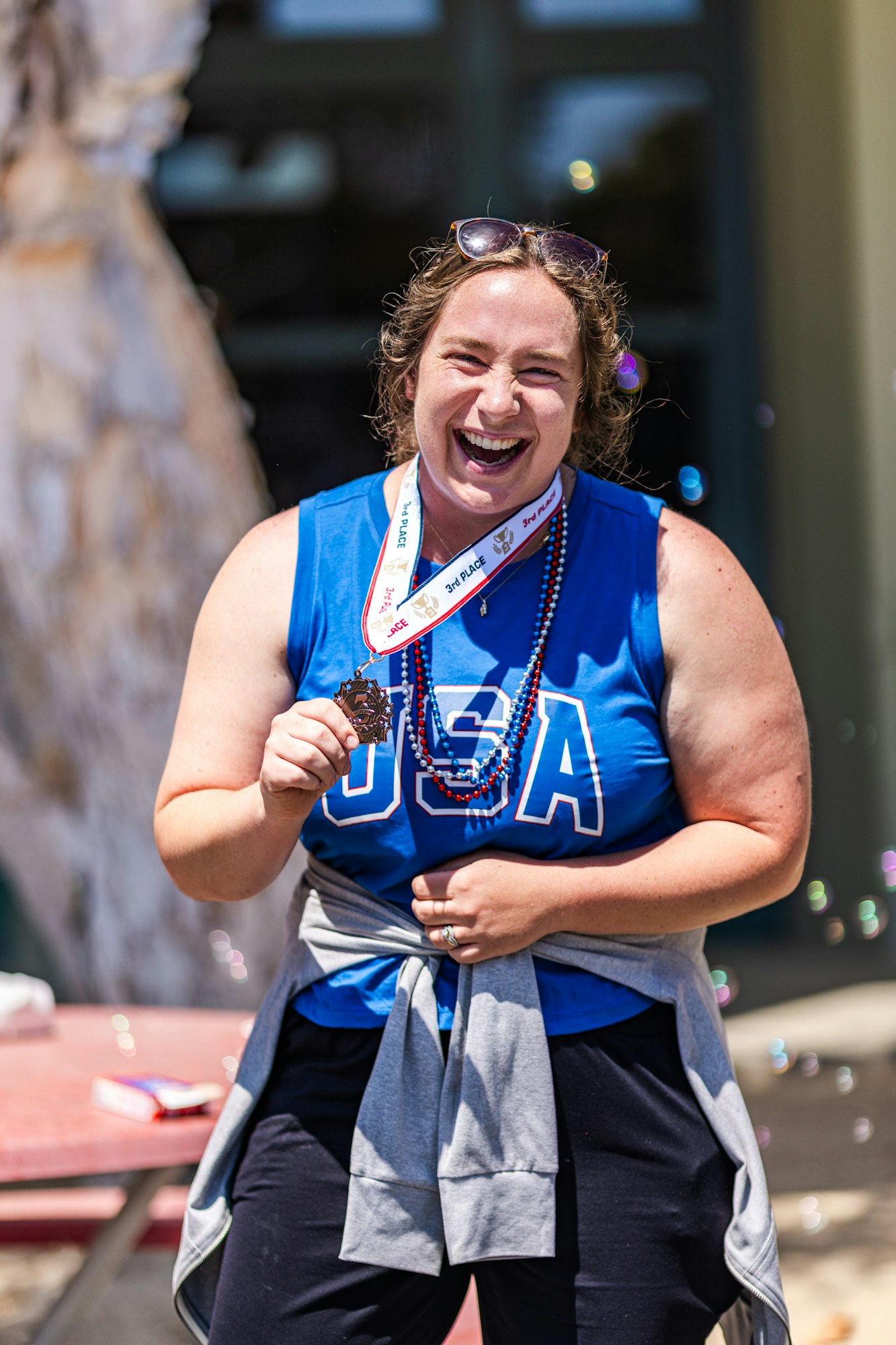 A smiling person in a blue "USA" shirt holds a medal labeled "3rd PLACE," standing outdoors with sunglasses on their head.
