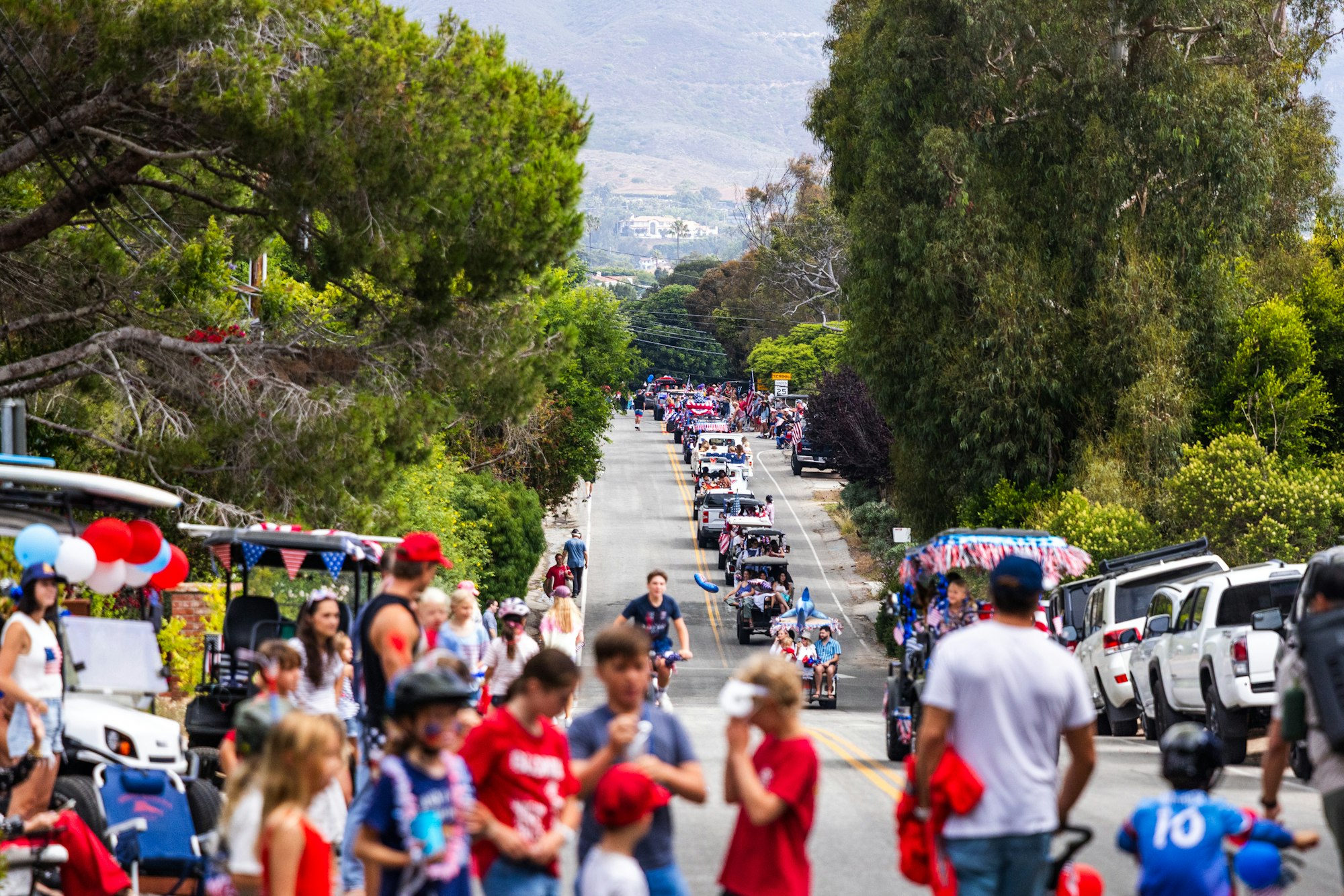 A festive street parade with people in red, white, and blue, featuring golf carts, balloons, and patriotic decorations.