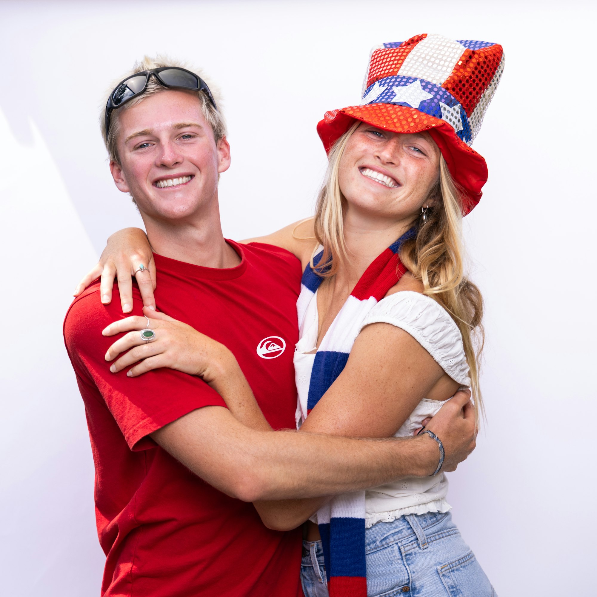 Two people smiling, one in a red shirt, the other wearing a red, white, and blue hat and scarf, embracing against a white background.