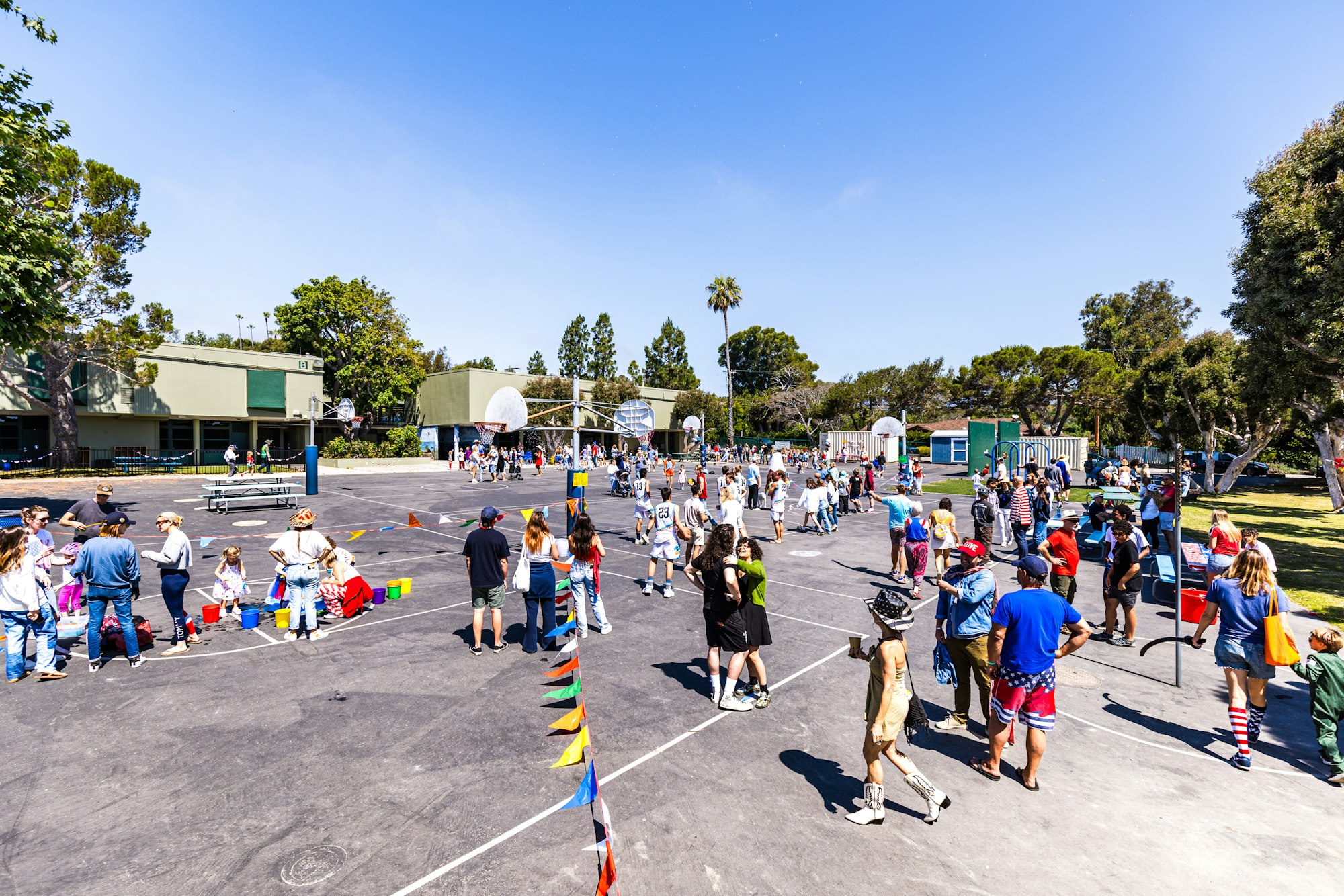 People gathered outdoors on a basketball court, engaged in activities, with colorful flags and trees in the background.