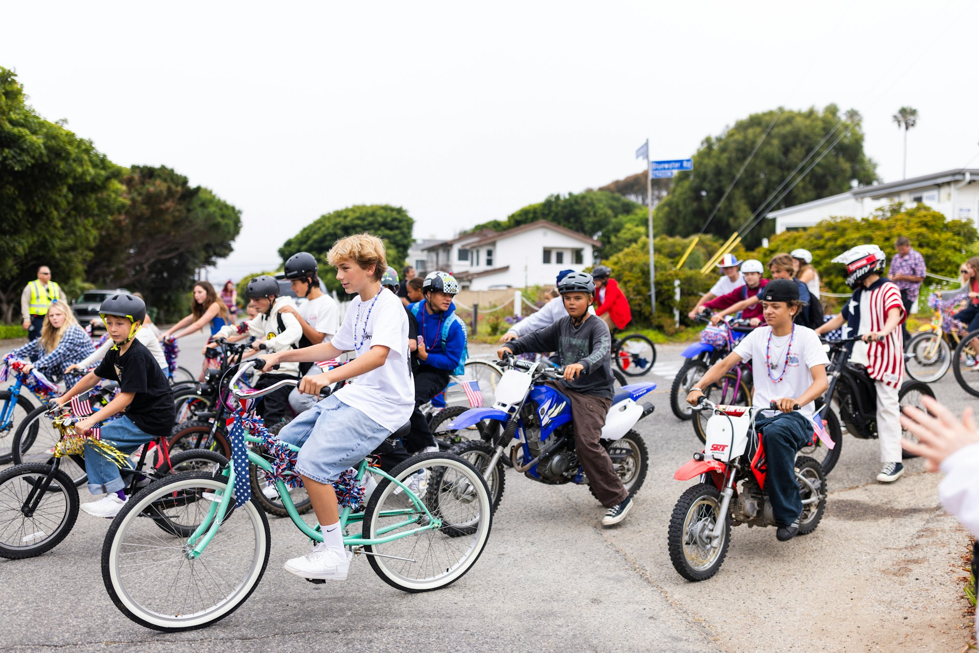A group of children riding decorated bicycles and dirt bikes on a street during a parade. They're wearing helmets and casual clothes.
