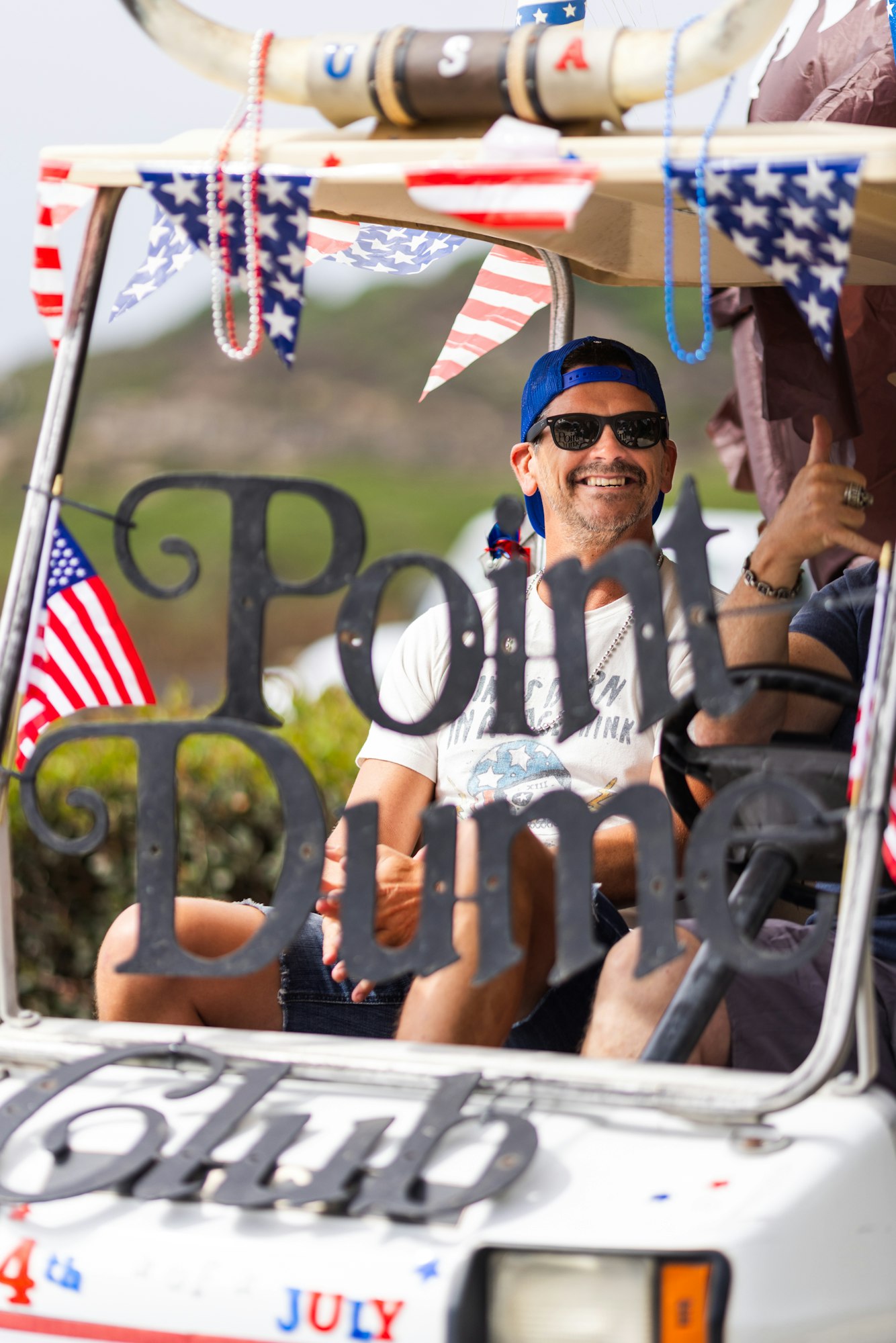 A man in sunglasses and a blue cap, smiling in a decorated golf cart with "Point Dume Club." American flags and bunting are visible.