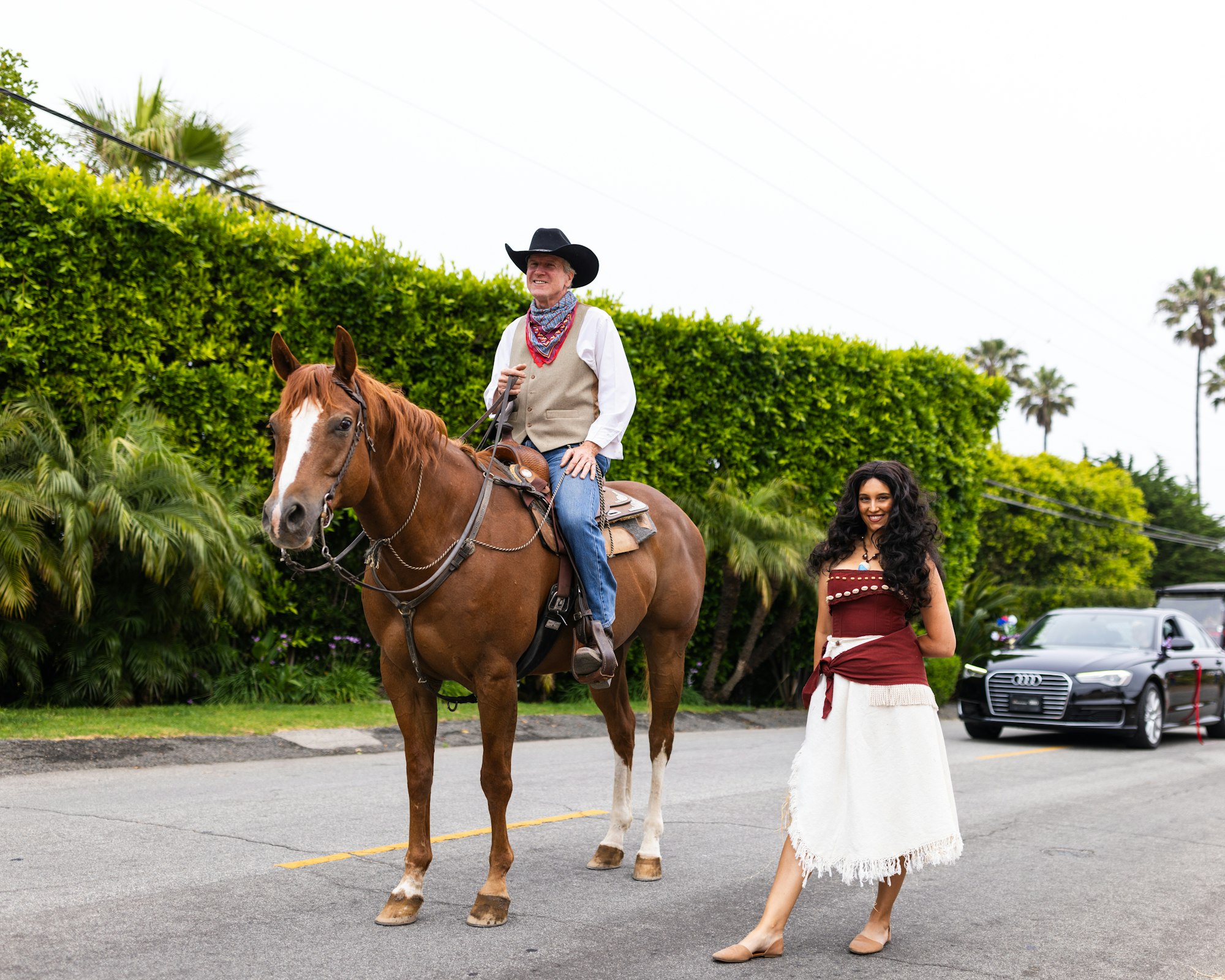 A man in cowboy attire rides a horse, while a woman in islander-inspired clothing stands beside them on a street.