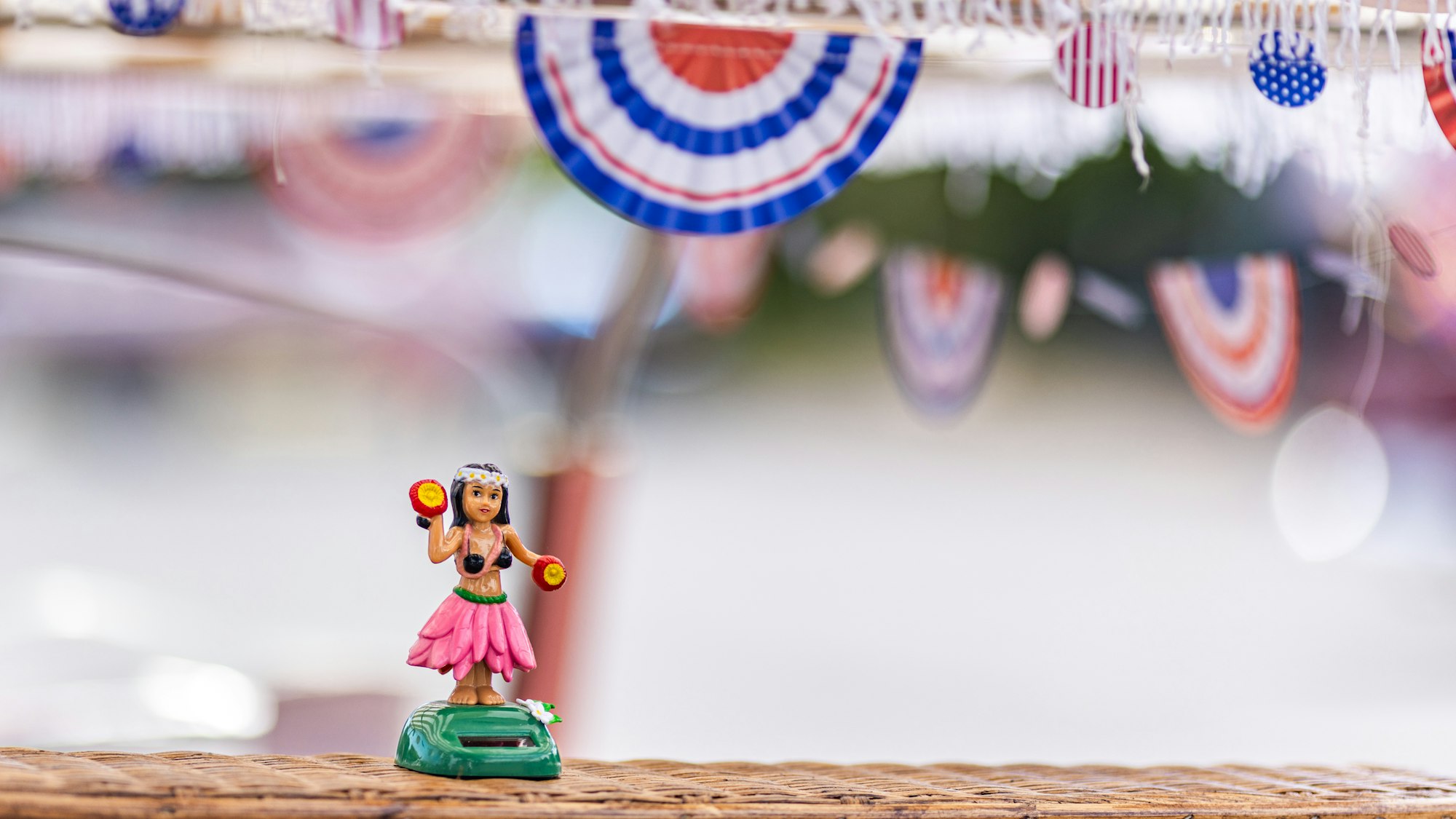 A small hula dancer figurine stands on a surface, with festive red, white, and blue decorations in the background.