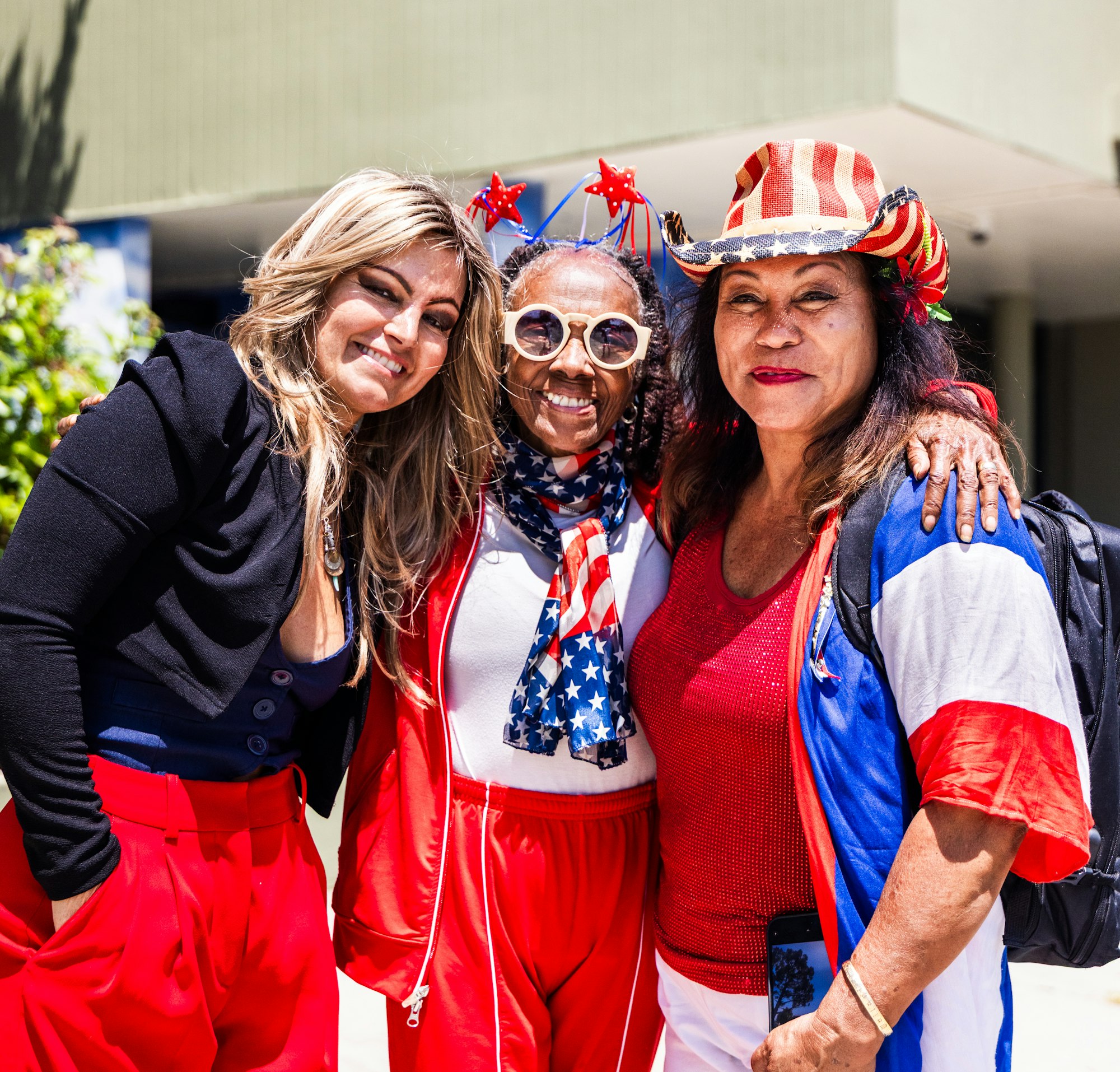 Three smiling women in patriotic red, white, and blue outfits, one wearing a star hat, outdoors.
