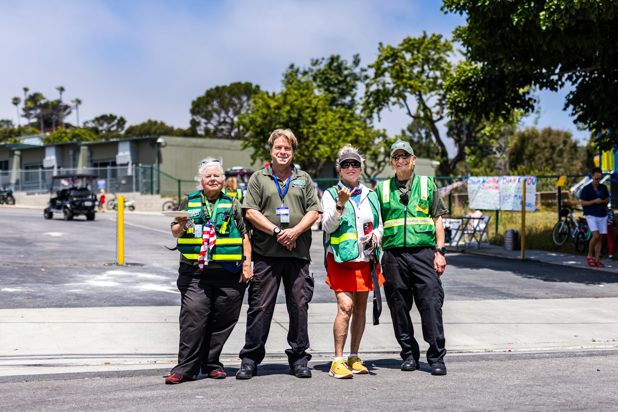 Four people in safety vests pose outdoors; background includes trees, pavement, and a building.