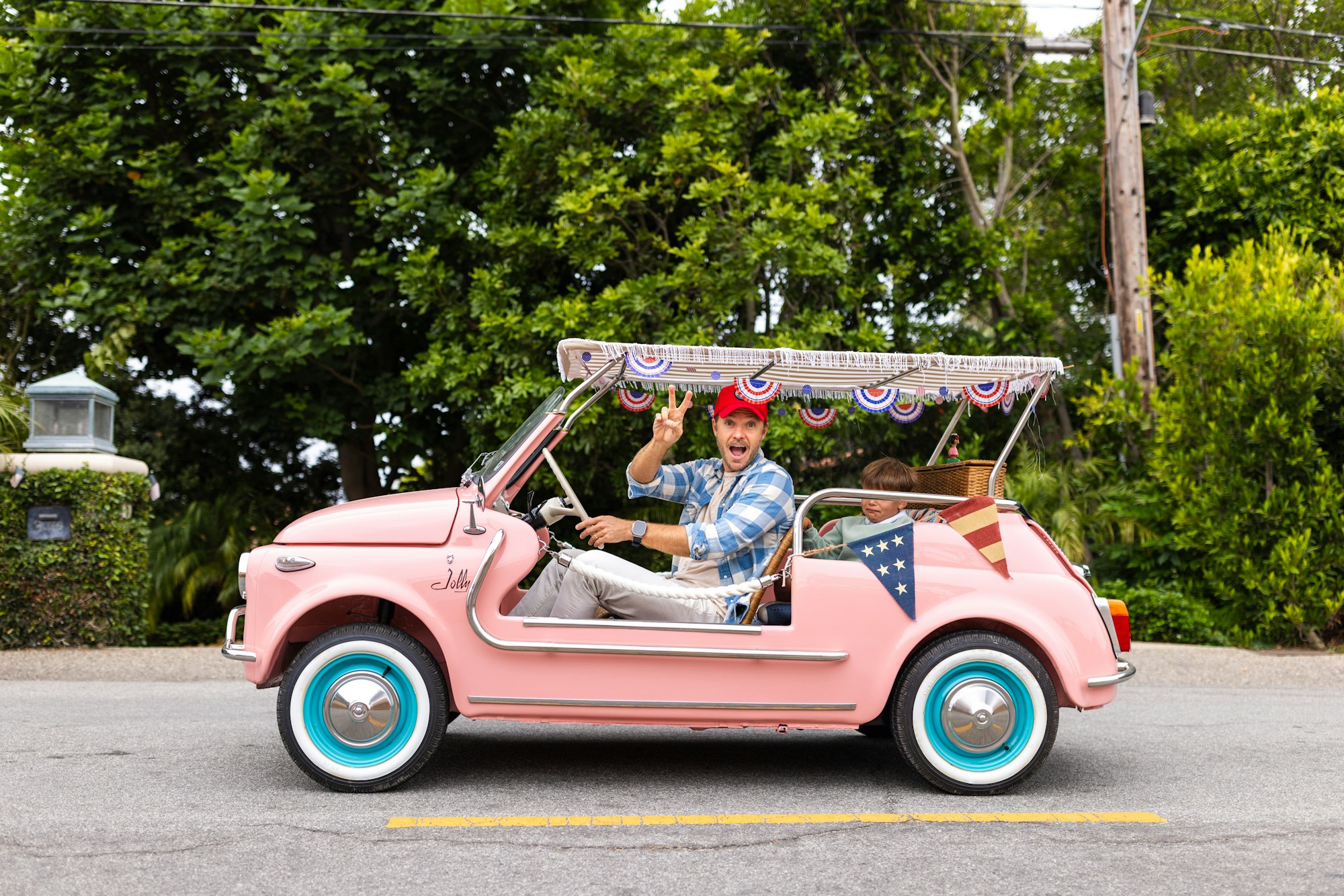 A man in a red cap and a child ride in a pink, open-top car decorated with patriotic bunting.