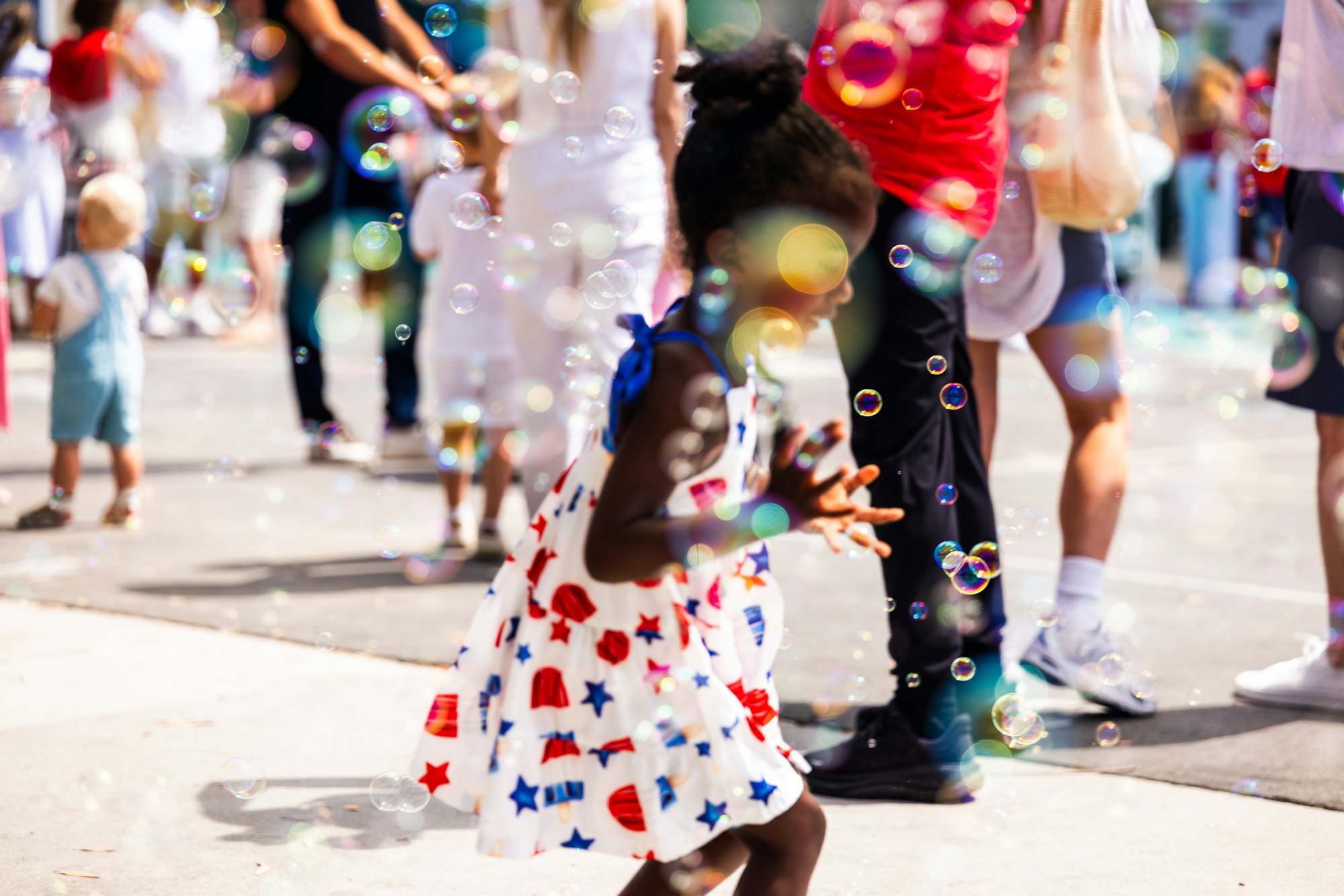 A child in a colorful dress plays with bubbles among a crowd on a sunny day.
