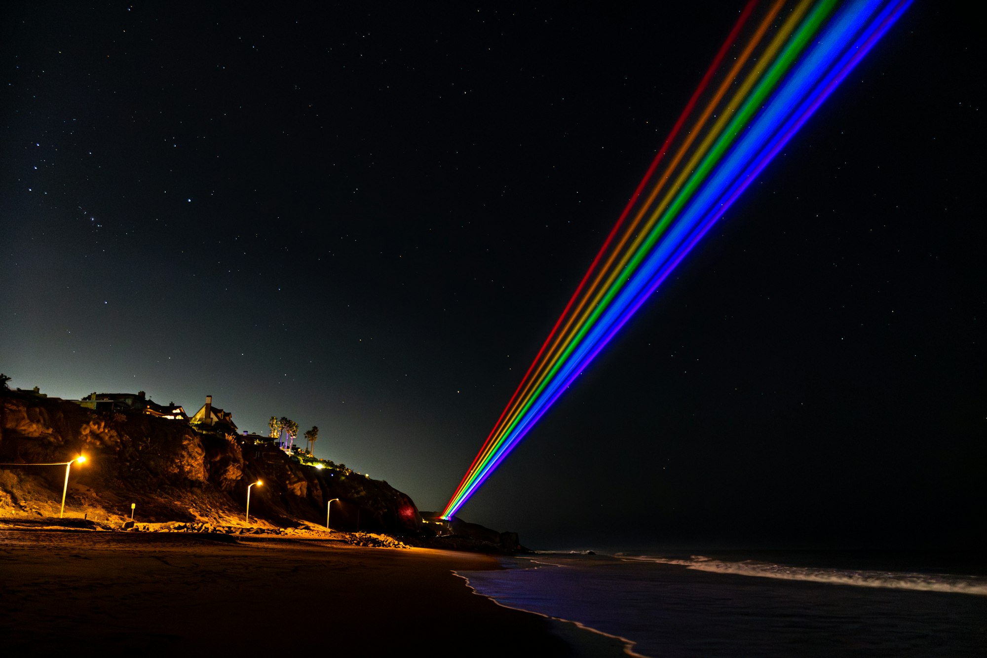 A dramatic night scene featuring bright rainbow lights beaming from a cliff, illuminating the beach under a starry sky.